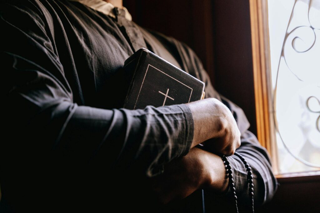 pexels-photo-8957588-8957588 A priest holding a Bible by a sunlit window, symbolizing faith and spirituality.